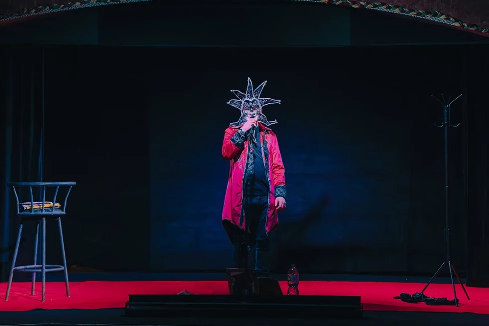 Un homme se produisant en spectacle. Il est debout et porte une longue veste rouge et un masque en forme de soleil sur le visage.