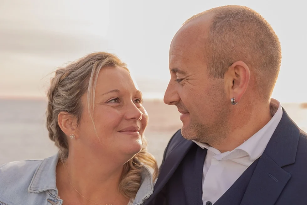 Couple de mariés souriants se regardant tendrement en extérieur, avec la mer en arrière-plan.