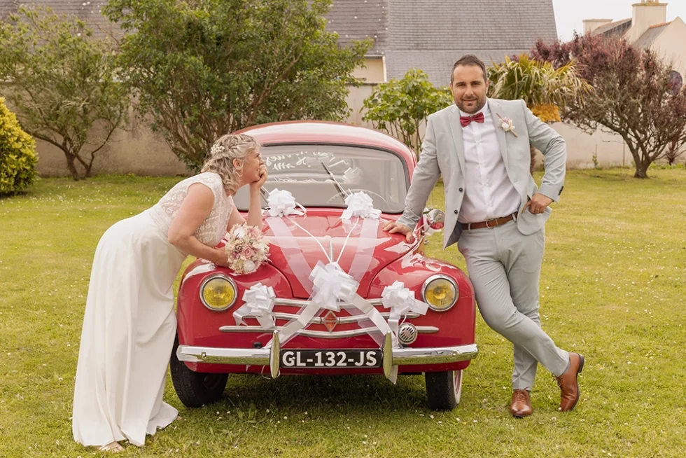 Couple de mariés en extérieur, la mariée en robe blanche adossée à une voiture ancienne rouge décorée pour le mariage, le marié en costume se tient debout de l'autre côté de la voiture.