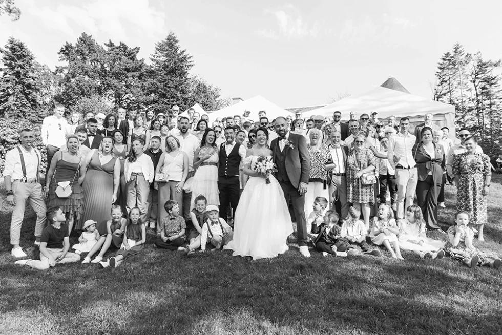 Photo de groupe d’un mariage en noir et blanc : les mariés au centre entourés des invités, prise en extérieur, avec des enfants assis au sol au premier plan.