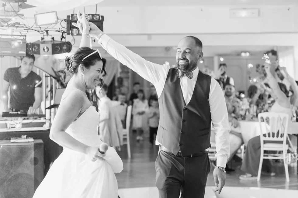 Photo en noir et blanc d’un couple de mariés dansant dans une salle de réception, la mariée tient un micro dans la main.