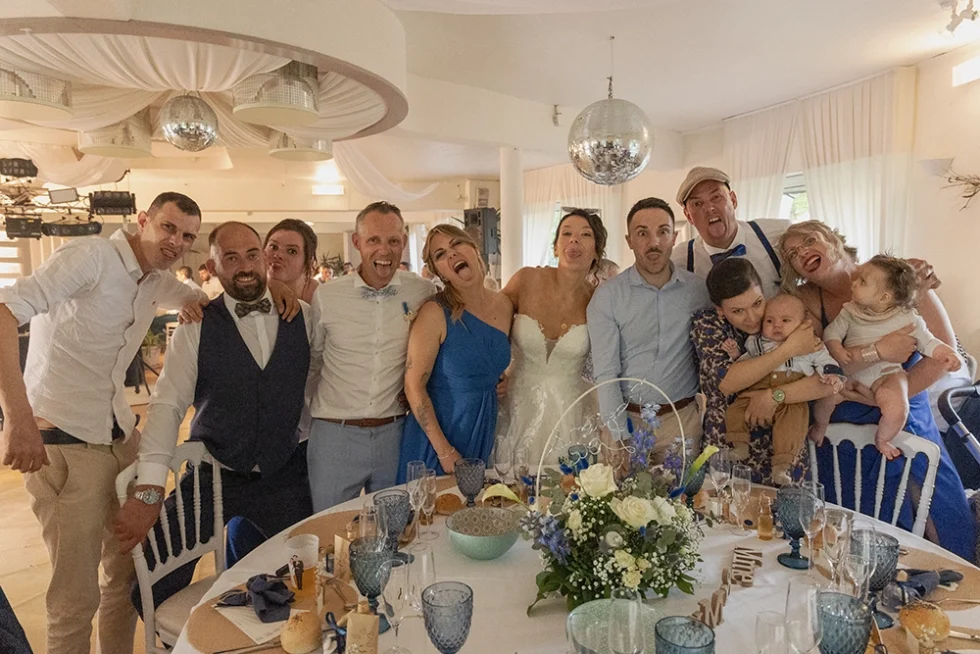 Famille réunie autour d’une table debout dans une salle de réception de mariage, faisant des grimaces pour la photo ; table décorée avec fleurs et vaisselle.