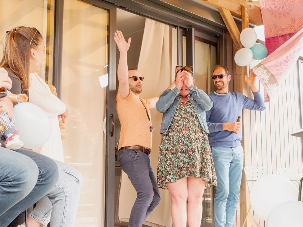 Jeune femme enceinte les yeux masqués par ses mains, entourée de deux hommes lors d’une baby shower, probablement surprise.