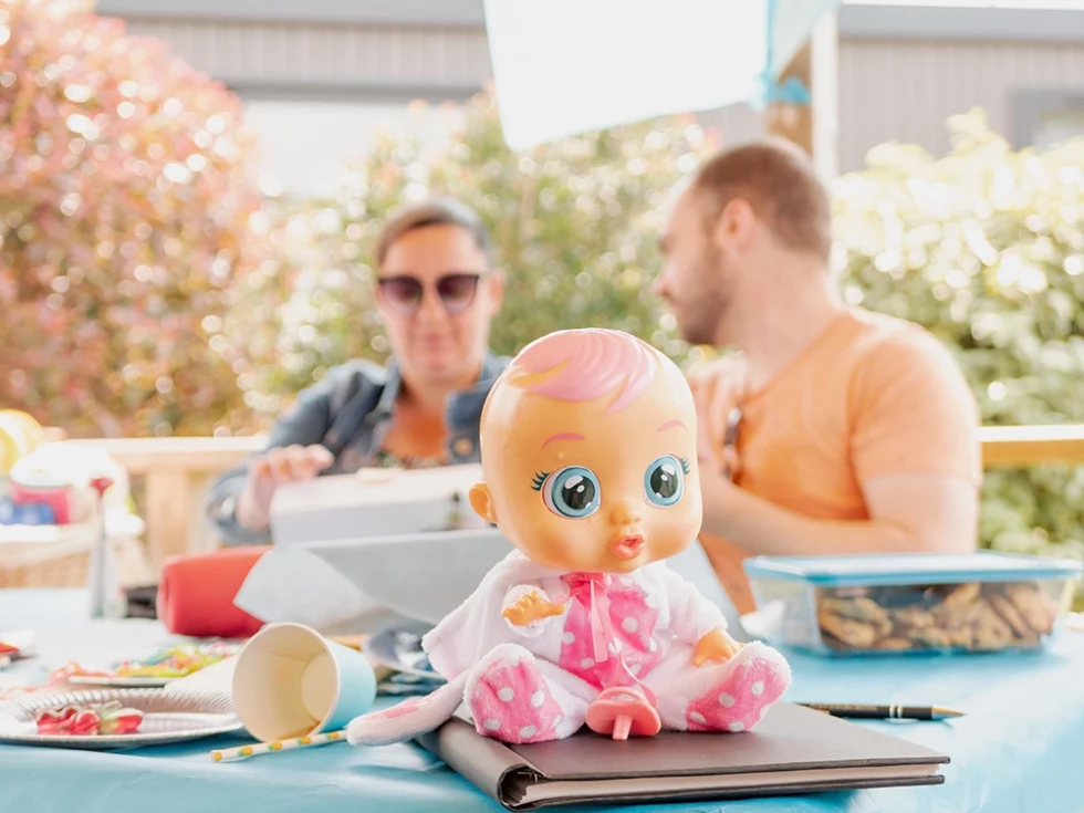 Lors d’une baby shower, une poupée rose avec tétine est posée sur une table au premier plan ; à l’arrière-plan, un homme et une femme discutent tranquillement.