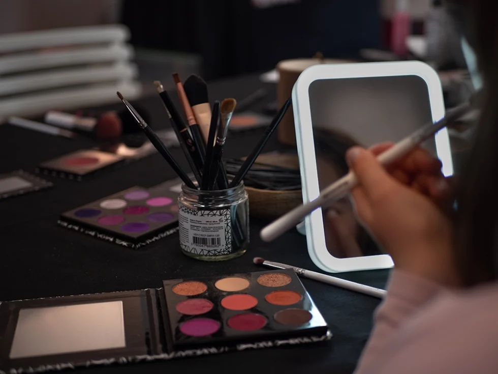 Femme qui se maquille devant un miroir. Elle tient un pinceau dans sa main. Des palettes de maquillage et un pot avec des pinceaux sont posés sur la table.