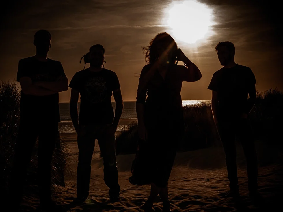 Groupe de quatre musiciens qui posent de nuit debout sur le sable. La mer est en arrière plan et le soleil se couche.