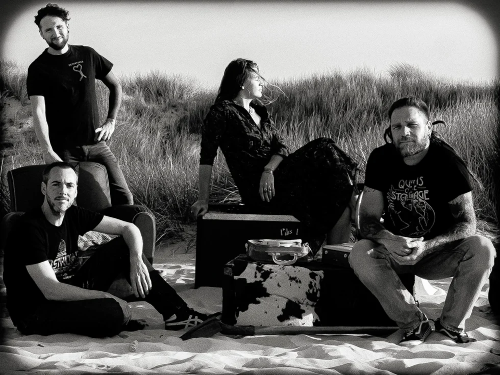 Groupe de quatre musiciens qui posent sur le sable avec de la végétation en arrière plan. Ils sont vêtus de noir. Il y a trois hommes et une femme.