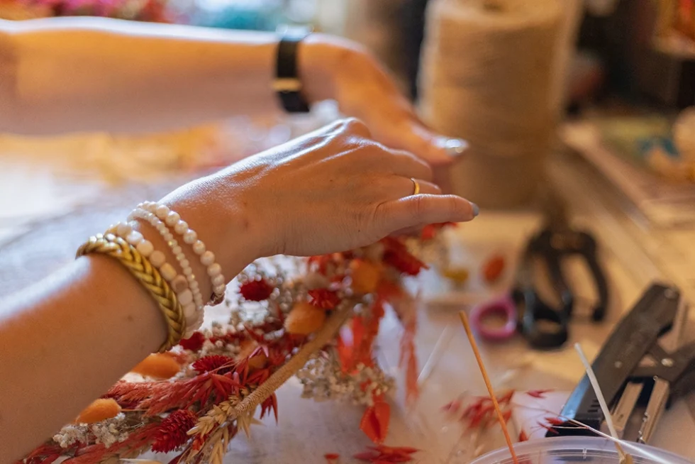 mains de femme ornées de bracelets au poignet en train de réaliser des compositions florales. Des ciseaux et une agrafeuse sont posées sur la table.