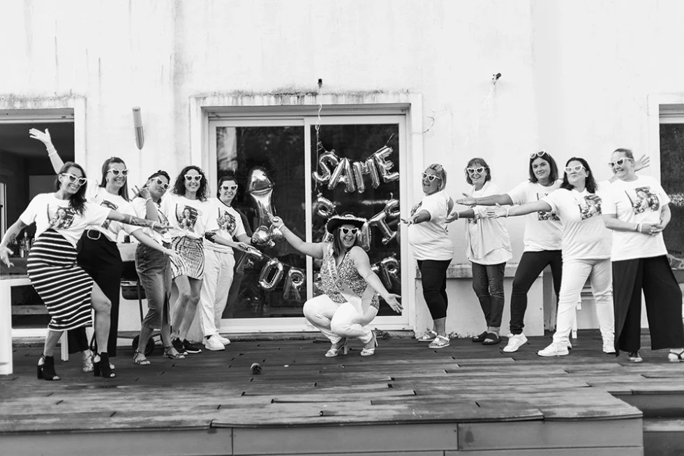 Photo en noir et blanc d'un enterrement de vie de jeune fille. La future mariée se trouve sur une terrasse devant une baie vitrée et elle est entourée de 10 amies.