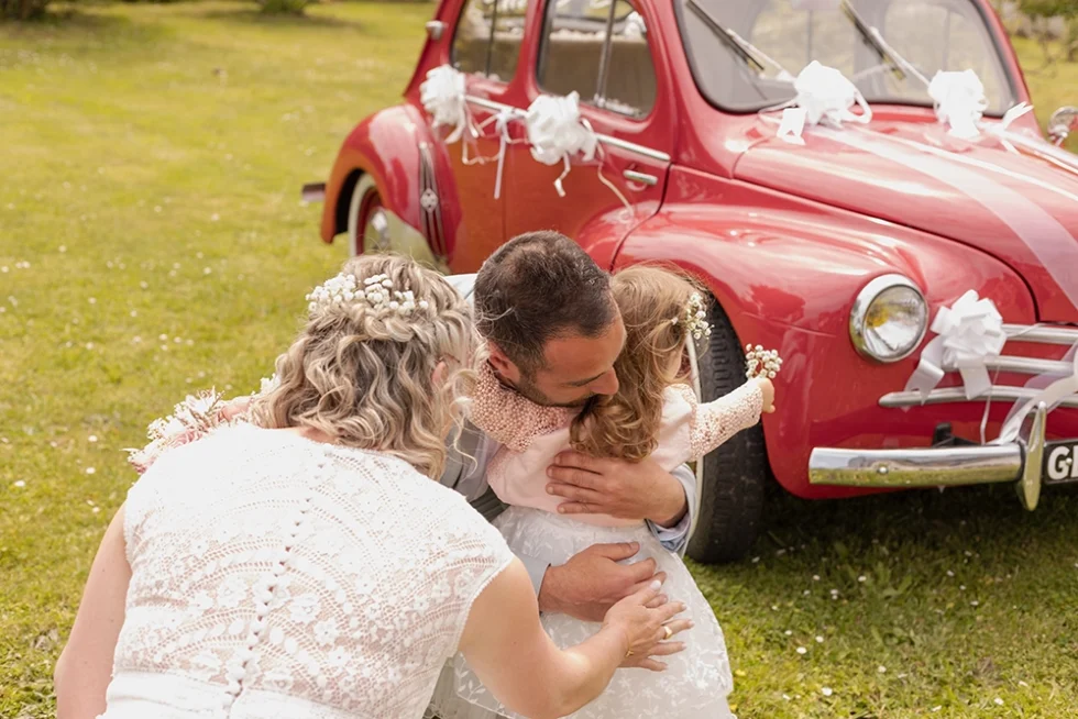 Devant une voiture ancienne rouge décorée de rubans blancs, un couple de mariés accroupis enlace leur petite fille, habillée comme sa maman en robe de mariée.