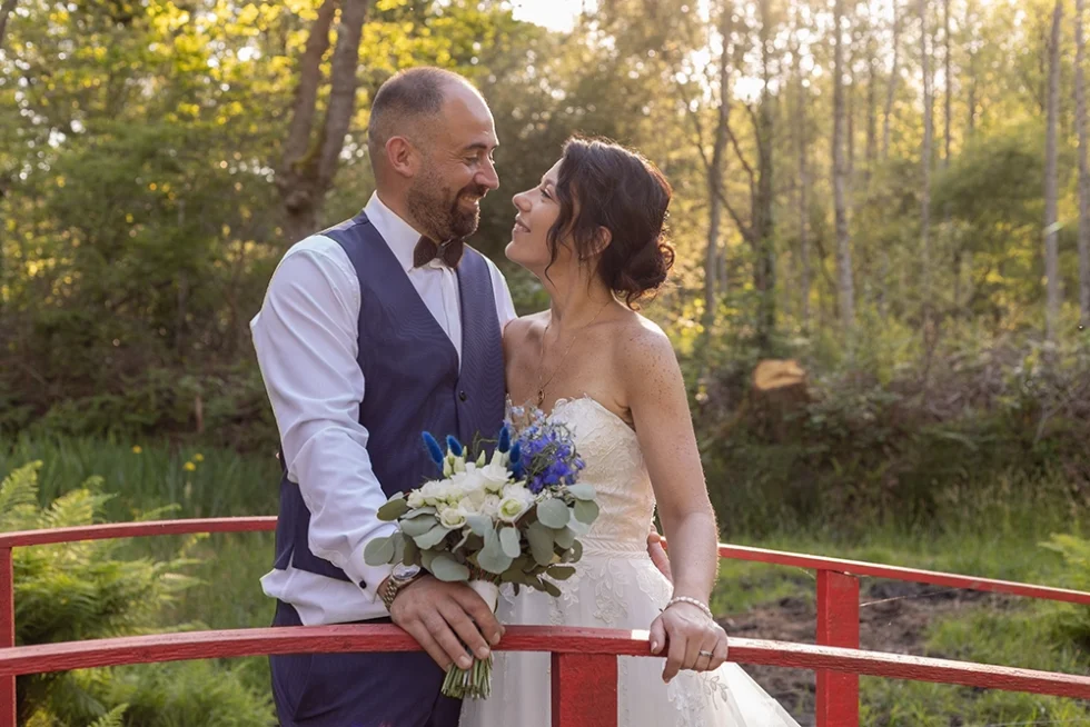 Mariés debout sur un petit pont aux rambardes rouges, se regardant avec tendresse ; le marié tient le bouquet de fleurs entre ses mains.