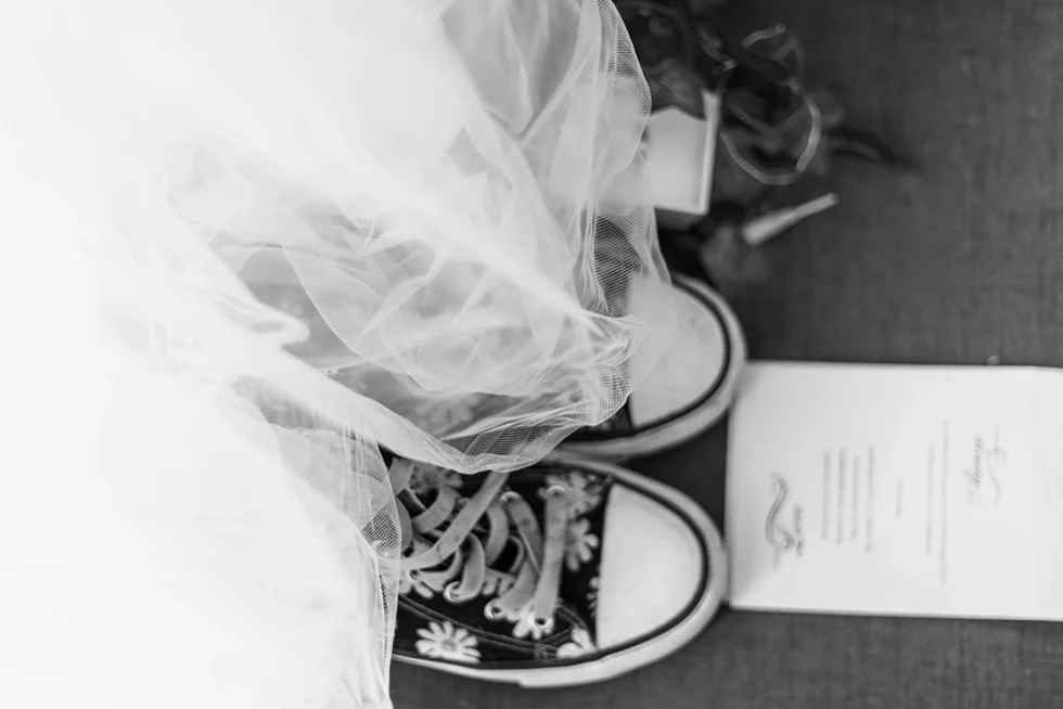 Photo en noir et blanc du bas d’une robe de mariée avec des baskets type Converse décorées de pâquerettes ; un faire-part de mariage est posé à ses pieds.