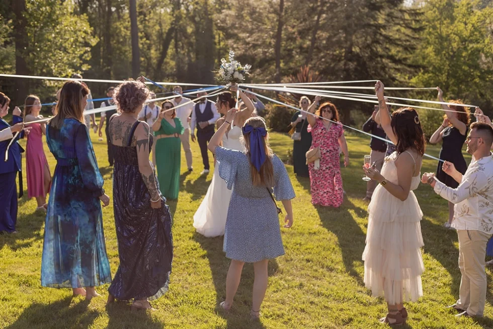 Mariée en robe blanche tenant son bouquet de fleurs levé dans les airs, entourée d’invités chacun relié au bouquet par un ruban.