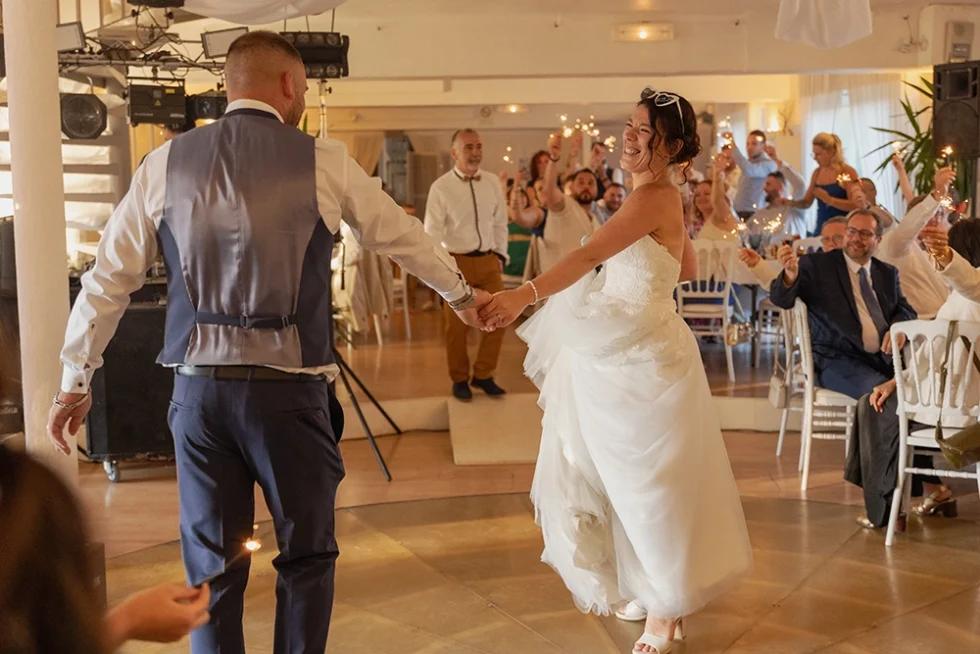 Couple de mariés dansant dans une salle de réception, entourés d’invités en arrière-plan tenant des bougies étincelantes.