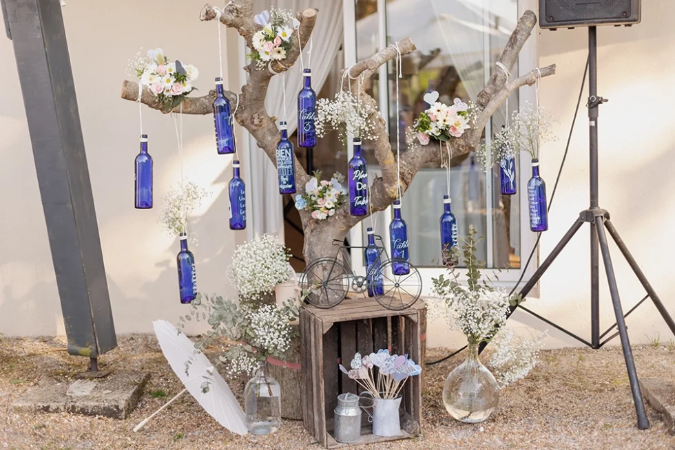 Scène de mariage : un arbre décoré de bouteilles bleues en verre avec un projecteur orienté vers une installation musicale en extérieur.