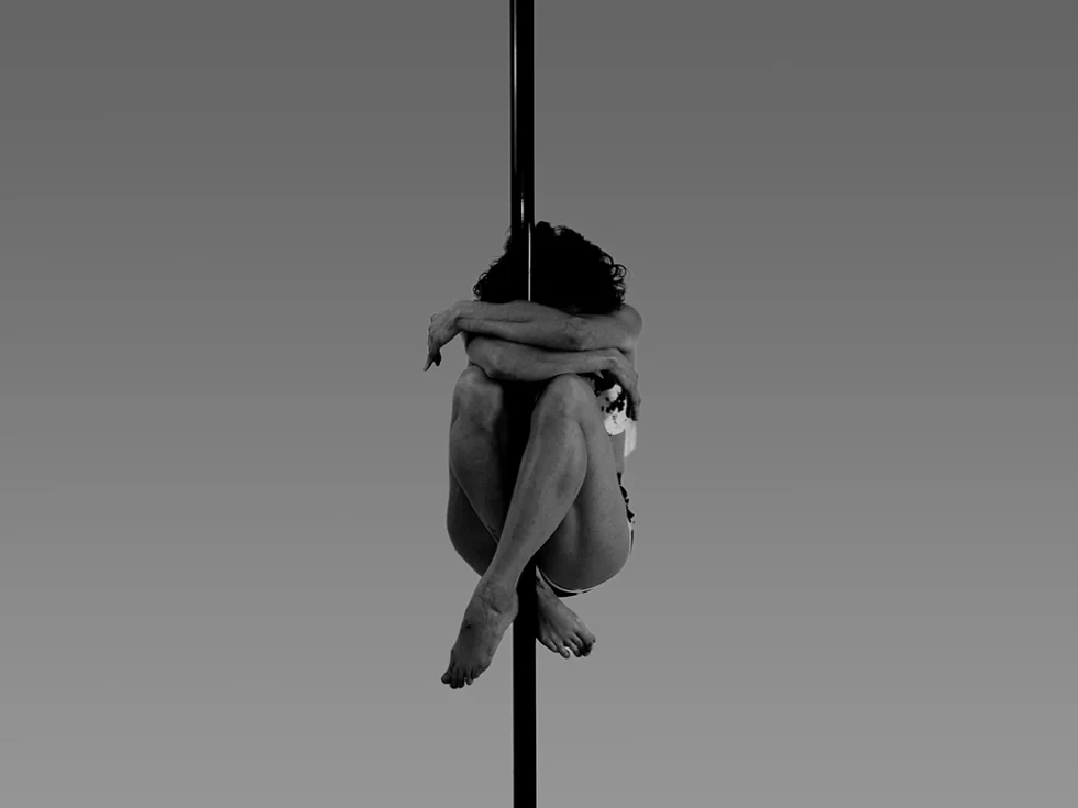 Photo noir et blanc d'une femme pratiquant des acrobaties autour d'une barre (pole dance) en tenue de sport.