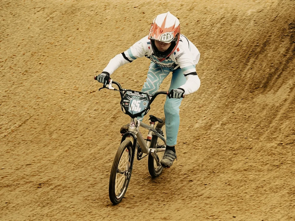 Jeune femme pratiquant du BMX. Elle est sur le vélo et porte un casque rouge et blanc avec une tenue de sport blanche et turquoise.