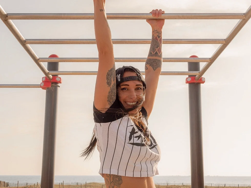Portrait d'une jeune femme qui réalise des acrobaties en extérieur. Elle porte une casquette noire à l'envers et un tshirt blanc à rayure noir. Elle est tatouée sur les bras.
