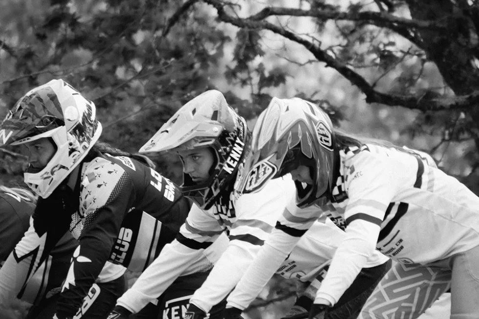 Photo en noir et blanc de trois sportifs au départ d'une course de BMX.