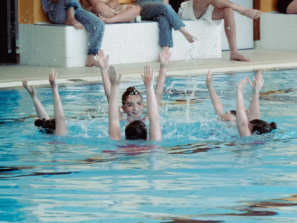 Groupe de cinq jeunes filles à la piscine qui réalise une chorégraphie artistique. Elles ont les bras levés.