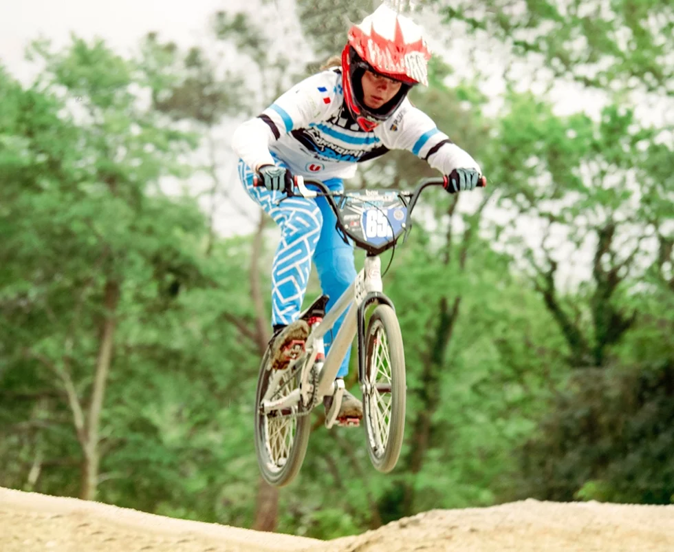 Jeune femme pratiquant du BMX. Elle est sur le vélo et porte un casque rouge et blanc avec une tenue de sport blanche et turquoise.