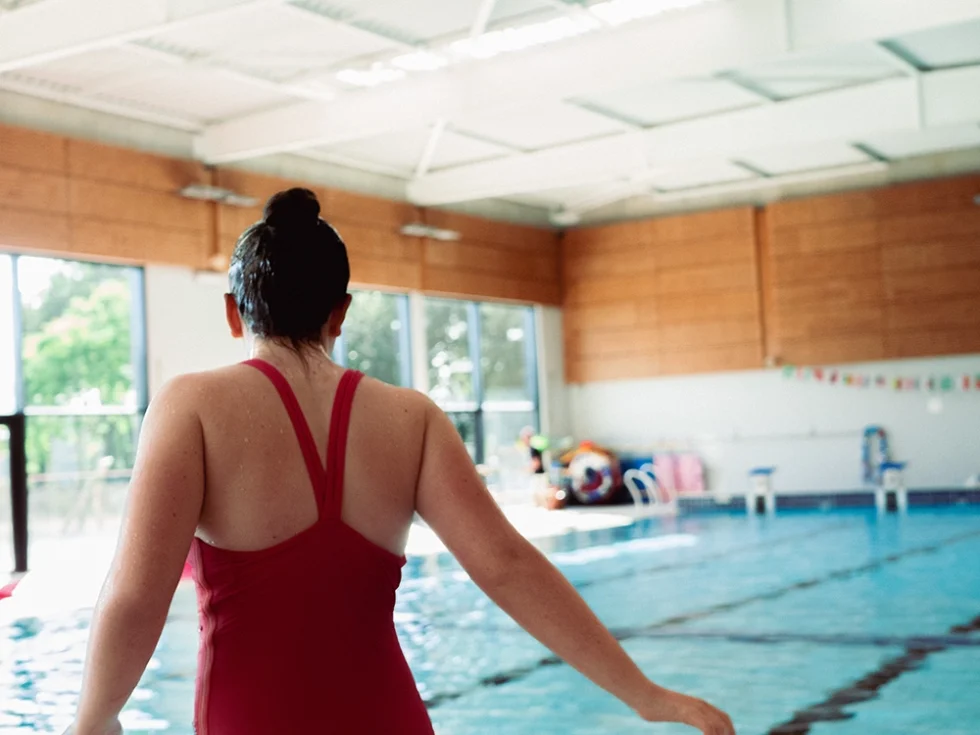 Jeune femme de dos à la piscine qui rentre dans l'eau. Elle porte un maillot une pièce rouge.